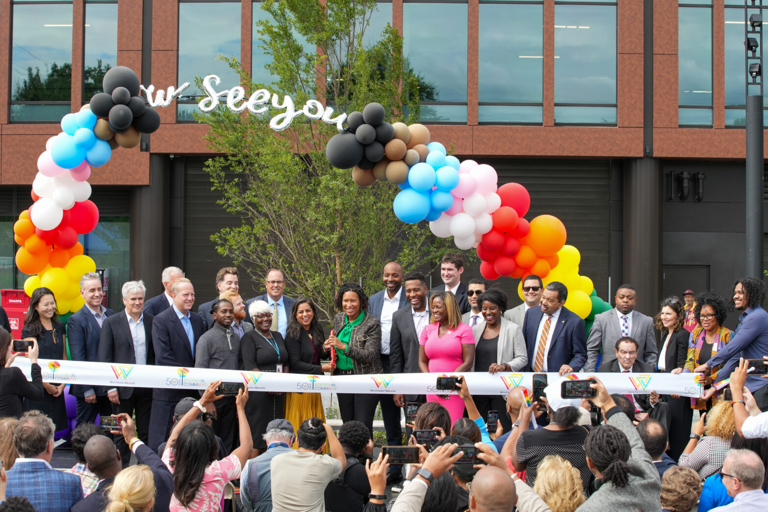 Group of dignitaries cut a ribbon onstage under a rainbow balloon arch with new medical building in background